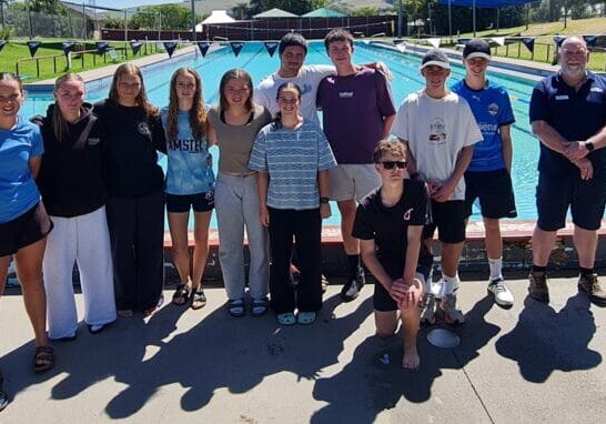 Group of teens and a coach posing poolside at a sunny outdoor swimming pool facility, with lane markers in background.